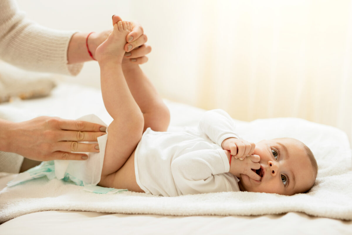 Baby boy lying on the bed with hands in the mouth while getting his diaper changed.